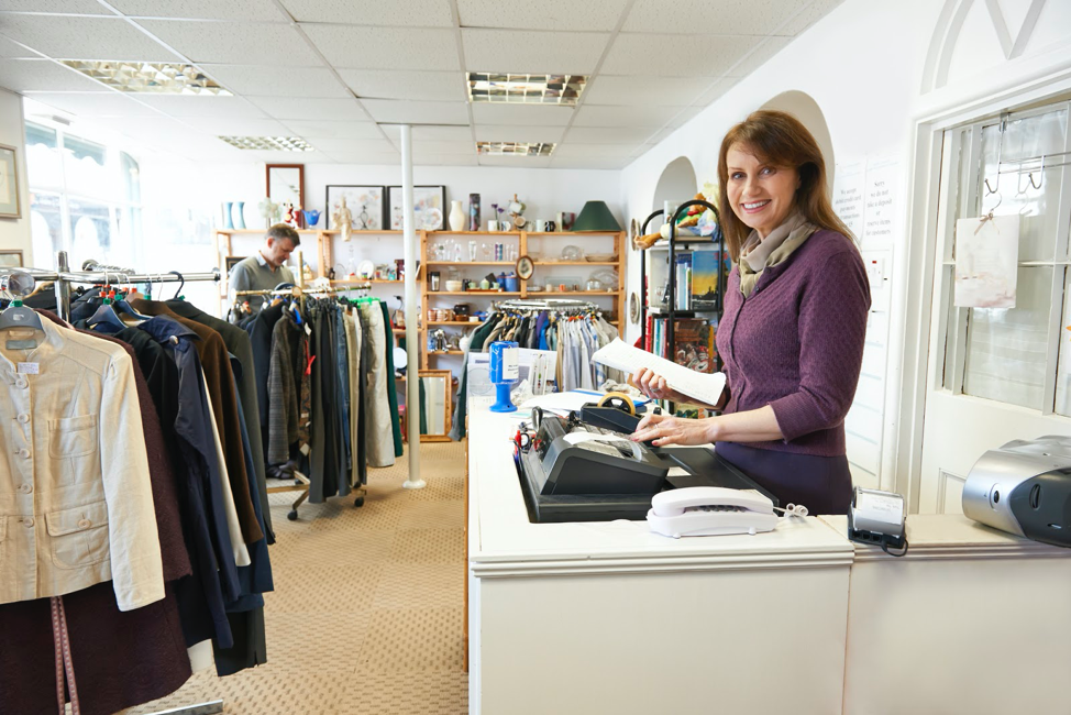 Woman in thrift store updating inventory at a POS register. Man shopping in the background.