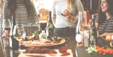 Three women standing around a table snacking and drinking wine