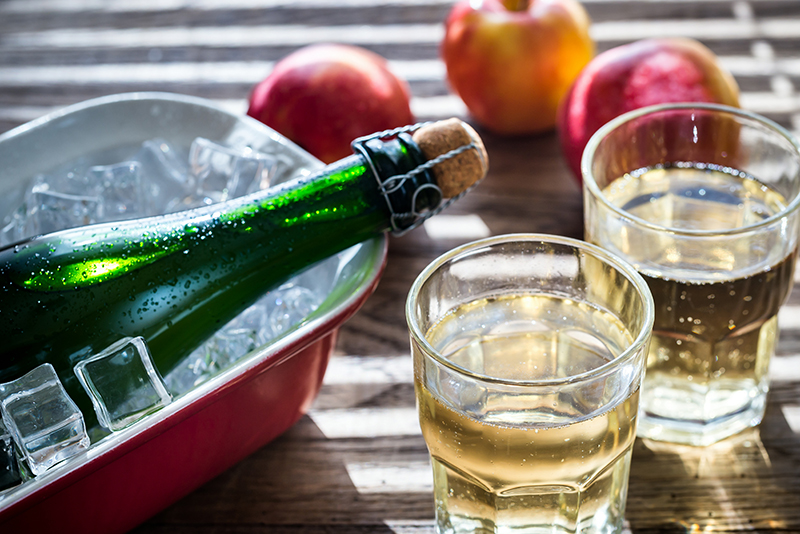 Cider in glasses with a bottle, ice and apples on a table.
