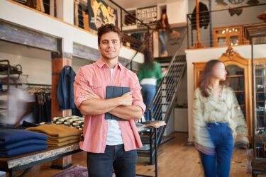 Man standing with tablet in the middle of a clothing store with customers around him.