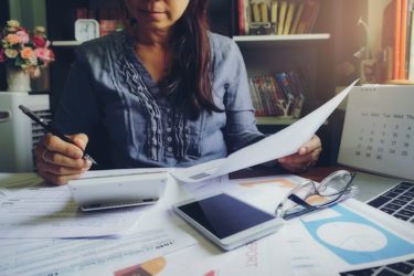 Woman sitting at a table with a bunch of papers, calculator, computer and glasses. She is working on filing her taxes