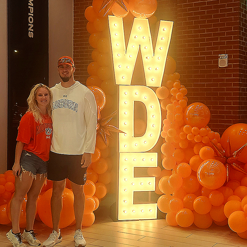 Abby and her fiancé wearing Auburn shirts standing in front of a giant WDE lighted sign.