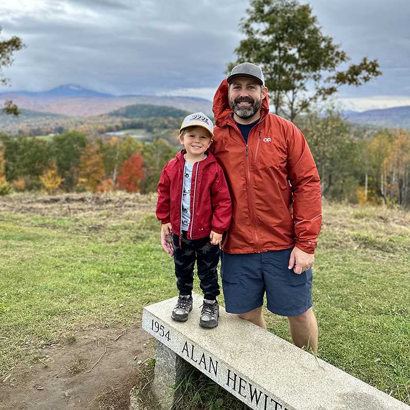 Ben with his son standing on a bench at a local hiking train in New Hampshire. The sky's are cloudy.
