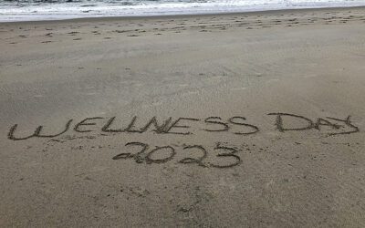 Wellness Day 2023 written in sand on the beach with the ocean in the background