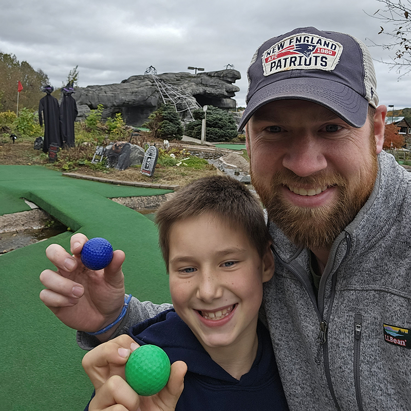 Dustin and his son holding golf balls with a mini golf course in the background.