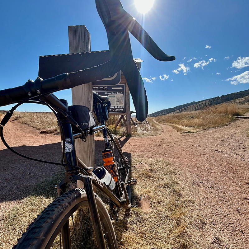 Gravel bike leaning against a post with the sun shining behind it and gravel trails in the background.