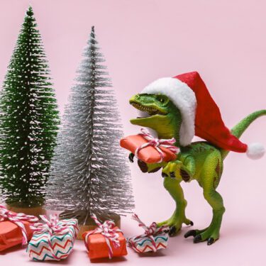 Funny plastic toy dinosaur wearing Santa Claus hat with present boxes on a pink background.