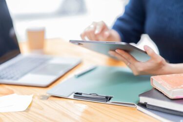 Woman reading reports on a tablet with papers and a laptop nearby.