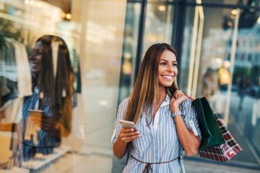 Woman holding shopping bags and her phone in front of a retail store.