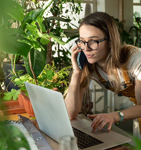 Woman uses laptop while talking with RCS support on smartphone. Small garden center business owner, entrepreneur receives expert trouble shooting advice online and over the phone.