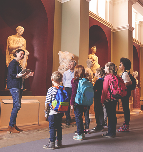 A group of kids looking at their teacher in front of a sculpture in a museum on a field trip.