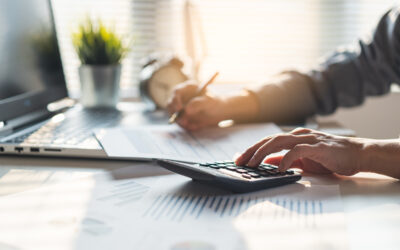 Man writing down tax information in a sheet of paper while looking at a computer and typing on a calculator.